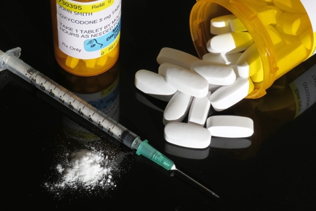 A white powder, needle, and prescription drugs sitting together on a tabletop.