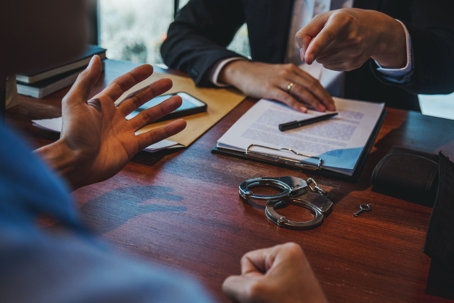 Two people sitting across from each other negotiating with paperwork and handcuffs sitting on a desk between them.